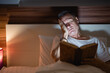 © Tj - Tired mature man struggling to stay awake while reading a book in bed at night, bathed in the warm glow of a bedside lamp, creating a cozy and peaceful atmosphere