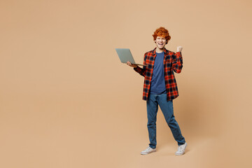 Full body young redhead IT man in red shirt blue t-shirt casual clothes hold use work on laptop pc computer do winner gesture isolated on plain pastel light beige background studio Lifestyle concept
