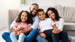 © Prostock-studio - Happy Loving Family. Portrait of cheerful African American man and woman sitting on the floor carpet in living room, posing for photo, embracing smiling boy and girl. Four positive black people