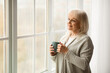 © Prostock-studio - Calm elderly woman standing near and looking out of window in living room, drinking tea, thinking or dreaming, smiling mature grandmother enjoying coffee relaxing at home, free copy space