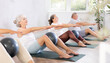 © JackF - Girl instructor does exercises together with elderly yoga practice visitors. People sit on karemat, pulled legs to themselves and clamped soft-ball in knees, perform simplified boat pose