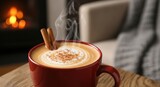 Steaming latte in a red mug with cinnamon sticks atop a swirl of milk. Background shows a fireplace and a cozy armchair draped with a blanket