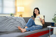 © Prathankarnpap - Smiling young woman sitting on sofa at home, holding smartphone and credit card for online shopping