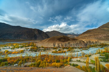 View of golden trees lining the glacial river contrasting with barren mountains under a dramatic sky, Yasin Valley, Gilgit Baltistan, Pakistan.