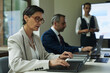 © DragonImages - Middle aged Caucasian woman typing on laptop with male colleague working beside her and young woman standing near digital presentation screen at business meeting