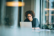 © Anastasiia Trembach - Focused woman working on laptop in modern office, wearing glasses with curly hair, showcasing concentration and professionalism.