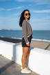 © sutulastock - Confident woman in striped sweater, black denim shorts, and sunglasses standing on a seaside promenade, smiling slightly with windblown hair, enjoying a sunny day near the ocean