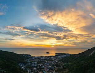 Wall Mural - A breathtaking sunset paints the sky in vivid hues of orange and blue over a coastal town, with lush green hills framing the scene. A distant hilltop structure adds a serene tranquil landscape.