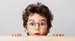 © ALLmost - A young boy with curly hair and round glasses peers over a wooden surface with a surprised expression.