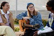 © Seventyfour - Group of multiethnic teenagers sitting outdoors, smiling and talking while young Caucasian girl with glasses and beanie holding acoustic guitar, enjoying casual conversation together