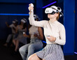 © JackF - Cheerful emotional young female in VR goggles sitting in virtual reality room with group of friends, manipulating objects or shooting at targets with gaming controllers in hands..