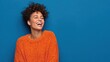 © Johannes - happy emotions concept positive dark skinned beautiful young woman laughs poisitively looks aside with carefree face expression wears casual orange sweater isolated over blue studio background no log