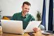 © Westend61 - Smiling man working from home using laptop holding documents