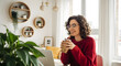 © mumtaz - Happy woman in red sweater enjoying coffee while working on laptop in bright modern home office with decorative shelves and plants