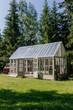© angel_nt - A lovely wooden greenhouse sits gracefully amidst tall evergreen trees, bathed in sunlight. The structure features glass panels and double doors, inviting nature inside