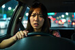 © Alex & M Studio - Stressed or scared young woman grips the steering wheel while driving through heavy city traffic at night, illuminated by the lights of other cars