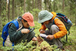 © Seventyfour - Two Black children crouching in forest examining moss and insects on fallen log using magnifying glasses, wearing backpacks and hats, exploring nature during outdoor adventure