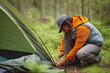 © Seventyfour - Black teenager girl crouching beside tent securing tent stake in forest setting, wearing cap and outdoor clothing, engaging in camping activity surrounded by greenery