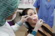 © Ekkasit A Siam - Asian adult female sitting in dental chair being examined by asian adultfemaledentist using dental mirror and scaler in clean white dental clinic with medical assistant nearby wearing blue uniform