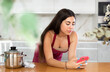 © JackF - ..Young brunette woman in apron reading message on smartphone in kitchen at home