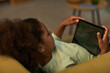 © DragonImages - Black girl lying on sofa holding digital tablet with both hands watching screen, relaxing at home, curly hair visible, casual setting, technology use by child