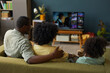© DragonImages - Black man, Black woman, and Black child sitting on sofa watching television together, man placing arm around woman, family spending leisure time in living room, backs facing camera