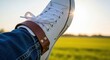 © MST - A person wearing white sneakers and denim jeans with a brown leather strap around the ankle enjoying the golden hour sunlight in a grassy field