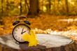© Julia - Alarm clock with a yellow maple leaf placed on a tree stump in a sunlit autumn forest with a blurred background. Symbolic image representing daylight saving time and fall season change