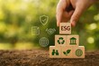© JH45 - A hand places a wooden block displaying the ESG acronym, amongst other sustainability icons, on a tiered stack of similar blocks positioned on earth.