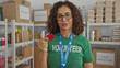 © Krakenimages.com - Hispanic woman in green volunteer uniform smiling indoors holding red heart symbol in charity room filled with donation items.