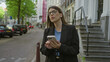 © Krakenimages.com - Woman holding smartphone on european street, wearing glasses and dark hair, standing among cars and buildings under green trees, exuding a professional vibe.