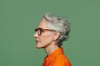 © Pavel - Mature woman with short gray hair and glasses poses against a solid green background, showcasing elegance and confidence in her expression