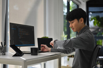 A Male software developer checking his smartwatch while working at a modern desk with code displayed on a monitor