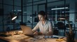 © patricia - A woman works at her laptop in an office lit by desk lamps at night