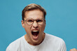 © SHOTPRIME STUDIO - Angry woman with short brown hair and glasses screaming against a plain blue background, expressing frustration or anger, studio portrait, emotional human face.