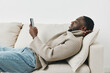© SHOTPRIME STUDIO - Young African man relaxing on a sofa while using a smartphone, casual outfit, cozy atmosphere, modern interior, light colors, and joyful expressions