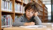 © Moopingz - A young boy with closed eyes rests his head on his hand, looking bored or tired while studying at a library desk with books around him.