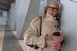 © Ivan Traimak - Stylish woman in neutral trench coat enjoying coffee outside modern building on a sunny day