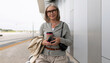 © Ivan Traimak - Senior woman holding a coffee cup and smartphone while waiting at an airport terminal on a cloudy day