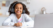 © Prostock-studio - Cute black school girl with wireless headset using laptop in kitchen, cheerfully smiling at camera, having online lesson during coronavirus pandemic. E-education, home schooling concept
