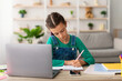 © Prostock-studio - Front portrait of focused girl sitting at table with laptop computer in living room at home, writing essay or making test exam, taking notes in textbook, doing homework, free copy space