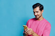 © SHOTPRIME STUDIO - Smiling man wearing pink t-shirt looks at his mobile phone, enjoying a moment of connection, captured against a solid blue background. Modern lifestyle concept.