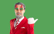 © Studio Romantic - Happy mature man dressed as Santa Claus, wearing funny reindeer glasses, pointing with thumb at copy space over green background, suggesting to write your advertisement or promo here