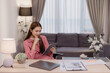 © Tj - Young Asian businesswoman working from home, sitting at her desk and using tablet with a pen in her other hand, with laptop and documents on the desk
