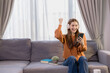 © Tj - Happy young woman enjoying music through wireless headphones, dancing joyfully while seated on a comfortable sofa in her cozy living room, embracing relaxation and vibrant energy