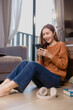© Tj - Happy young woman sitting on the floor of her cozy living room, using her smartphone while smiling and enjoying her time at home in casual, comfortable clothing