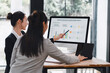 © kenchiro168 - Young businesswoman presenting quarterly growth chart on desktop monitor to colleague in modern office, collaborative meeting with tablet and documents, focused professional discussion