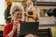 © La Famiglia - Smiling student working on laptop and drinking coffee outdoors on campus