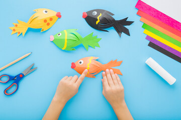 Wall Mural - Little child boy hands creating and gluing fish shapes. Scissors, glue stick, pencil and colorful application paper on blue table background. Pastel color. Closeup. Point of view shot. Top down view.