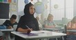 © Framestock - Muslim Primary School Girl Wearing Hijab Sitting at the Desk, Doing Task in Notebook During Lesson or Lecture, Writing School Test. Group of Young Students Studying in Modern Multicultural Classroom.